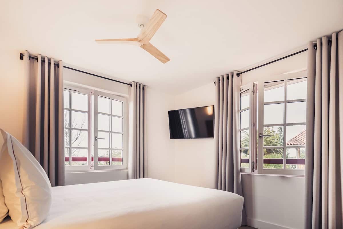 Une chambre d’hôtel avec un lit, un ventilateur de plafond, un téléviseur fixé au mur et de grandes fenêtres avec des rideaux.