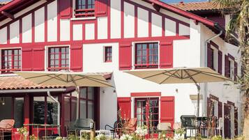 A two-story building with red shutters and white walls, featuring outdoor seating with umbrellas.