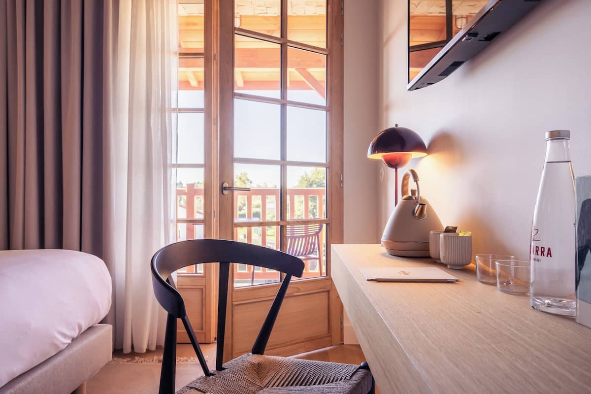 Une chambre d’hôtel avec un lit, une chaise, un bureau avec une lampe, une bouteille et une fenêtre donnant sur une terrasse en bois.
