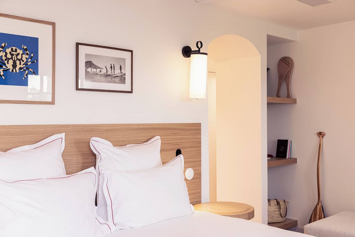 Une chambre d’hôtel avec une tête de lit en bois, un lit avec une literie blanche, un tableau encadré au mur, un luminaire fixé au mur et une petite étagère en bois sur laquelle se trouvent un livre et un sac à main.