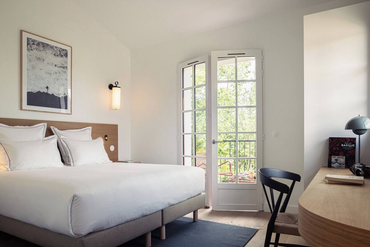 Une chambre d’hôtel comprenant un lit, un bureau, une chaise, un tableau encadré et une fenêtre donnant sur un paysage verdoyant.
