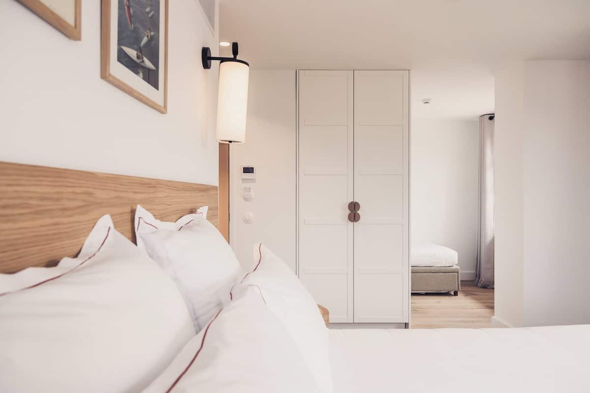 Une chambre d’hôtel avec une tête de lit en bois, une grande armoire, une table de chevet avec une lampe et un lit avec du linge de lit blanc.
