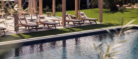 A poolside area with lounge chairs and umbrellas, surrounded by a wooden pergola and a well-maintained lawn.