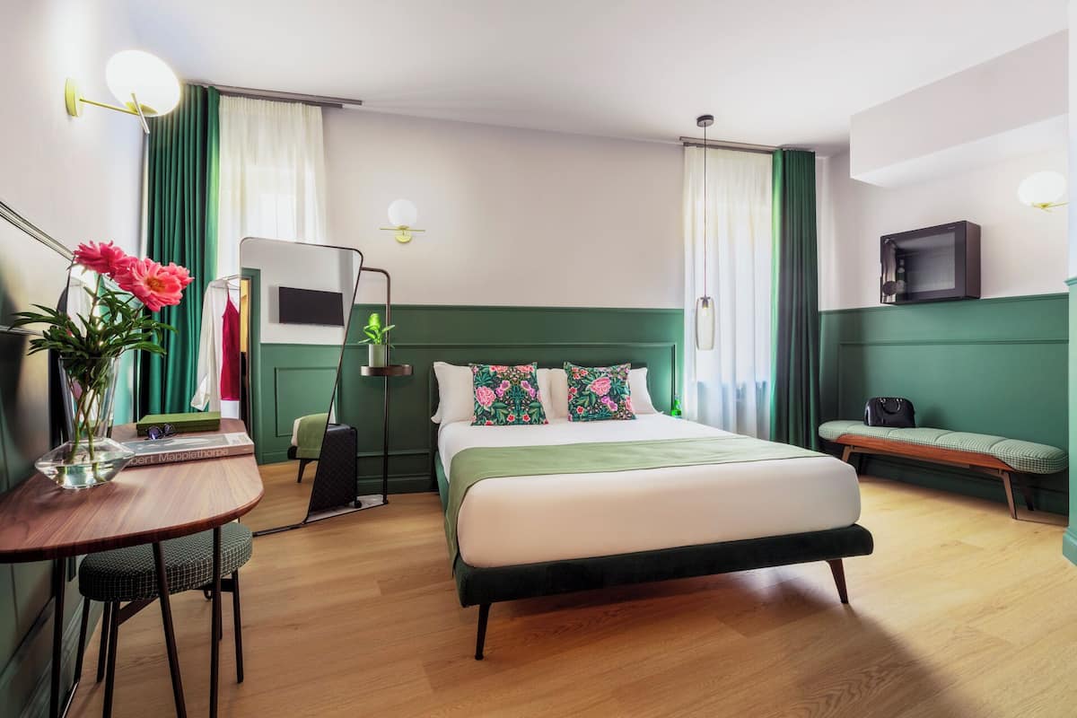 A hotel room with a large bed, a desk with a chair, a bench, and a small table with a vase of flowers.