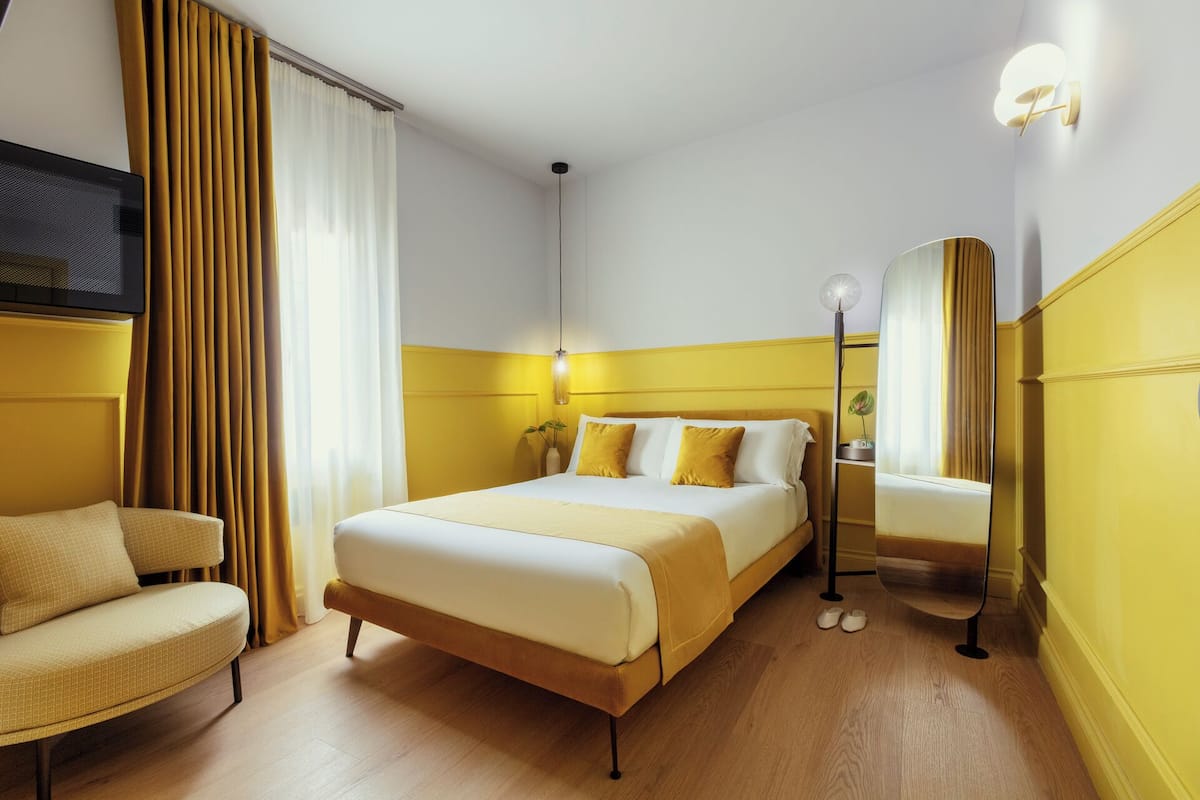 A hotel room with a bed, a chair, a bedside table, a lamp, and a mirror.