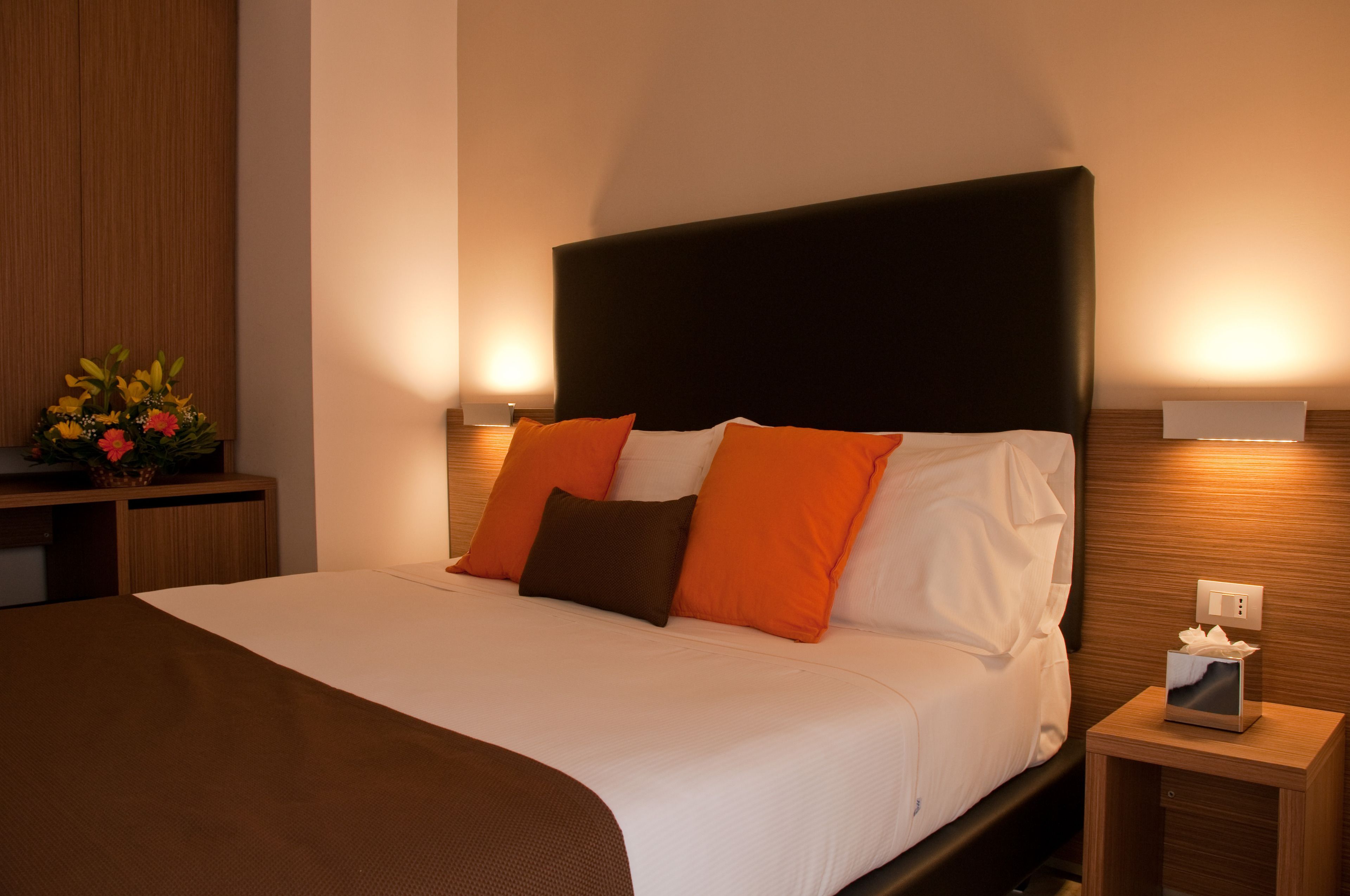 A neatly made bed with white and orange bedding, a dark headboard, and a bedside table with a tissue box and a small picture frame.