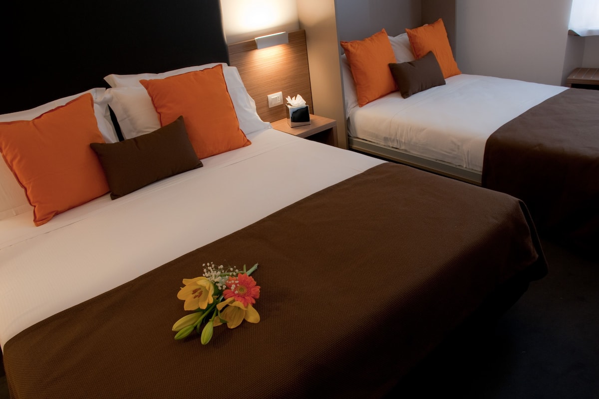 Two double beds with orange and brown bedding, a nightstand with a tissue box, and a floral arrangement on the bed.