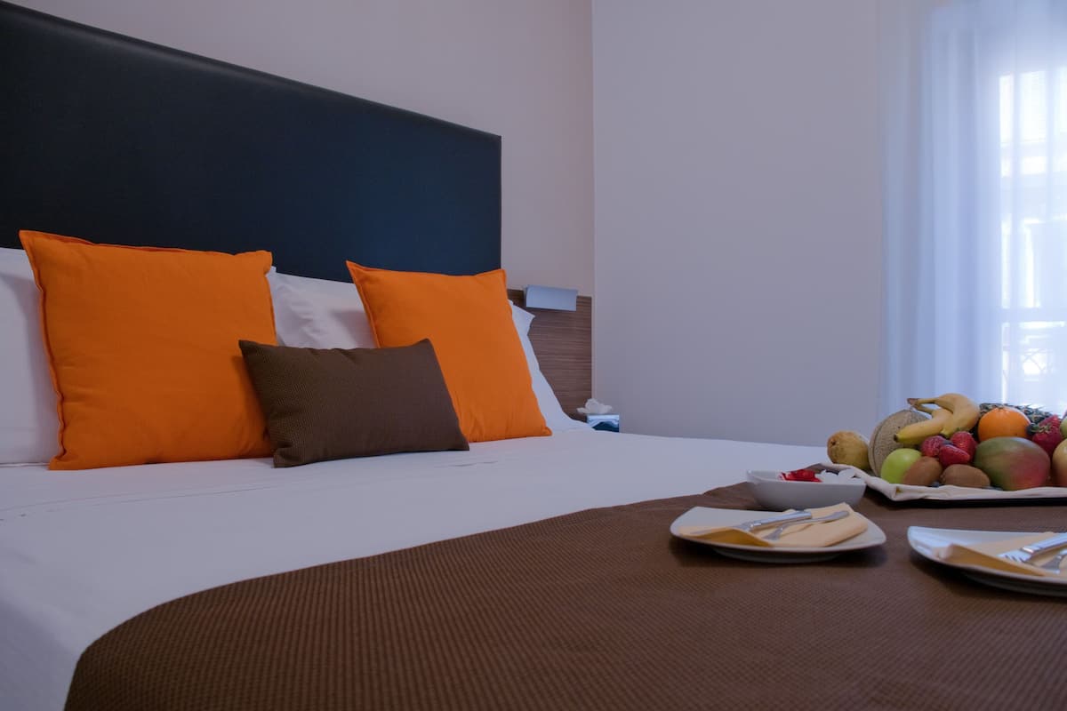 A hotel room with a neatly made bed, orange and brown pillows, a fruit basket on the nightstand, and a breakfast tray on the table.