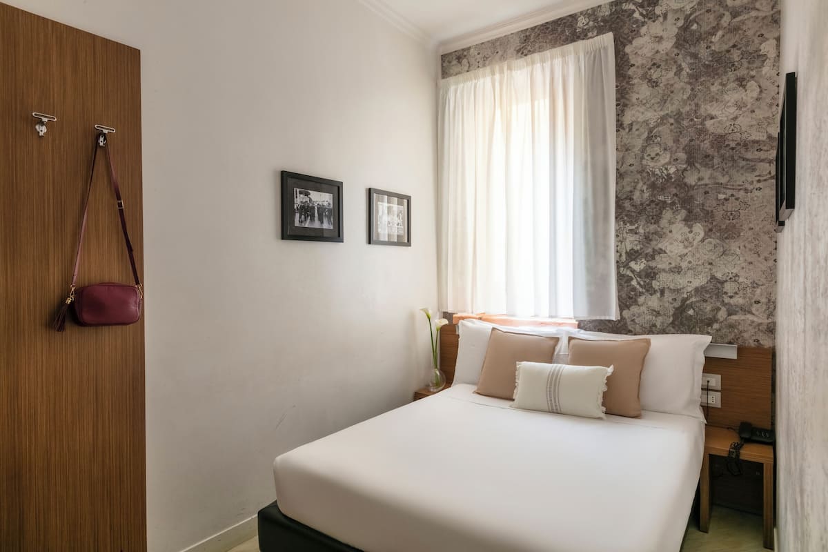 A neatly arranged hotel room with a bed, a wooden wardrobe, a small table with a lamp, and a wall-mounted TV.
