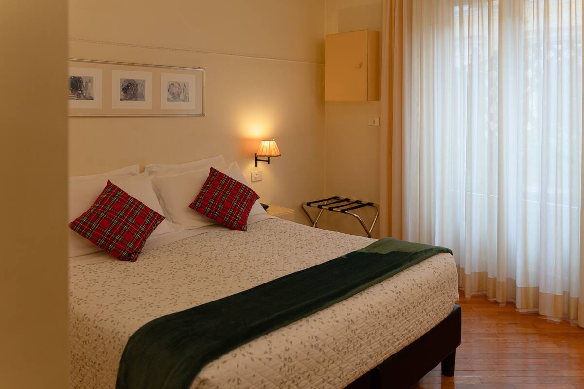 Un lit bien fait, recouvert d’une couvre-lit à motifs, avec deux oreillers à carreaux, un plaid vert, une lampe fixée au mur et une fenêtre ornée de rideaux légers.