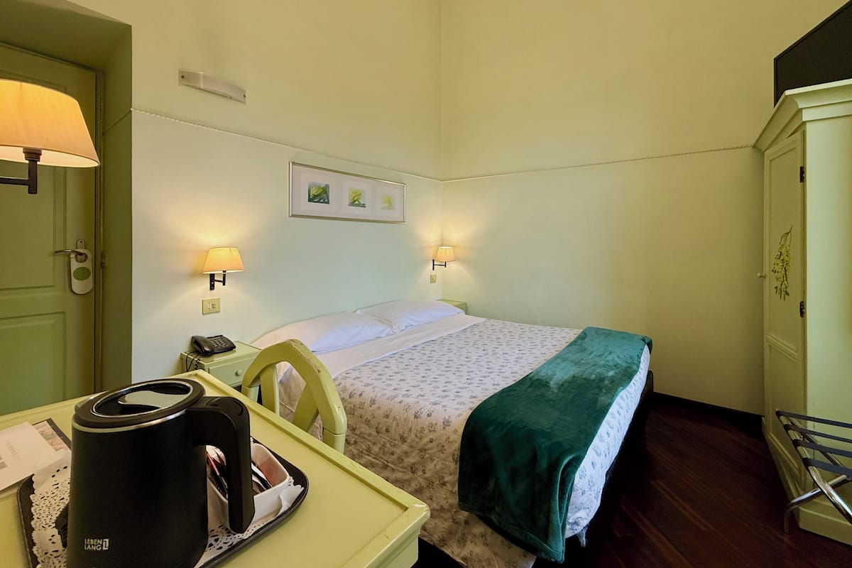Une chambre d’hôtel avec un lit, un bureau avec une bouilloire, une chaise et une couverture verte.