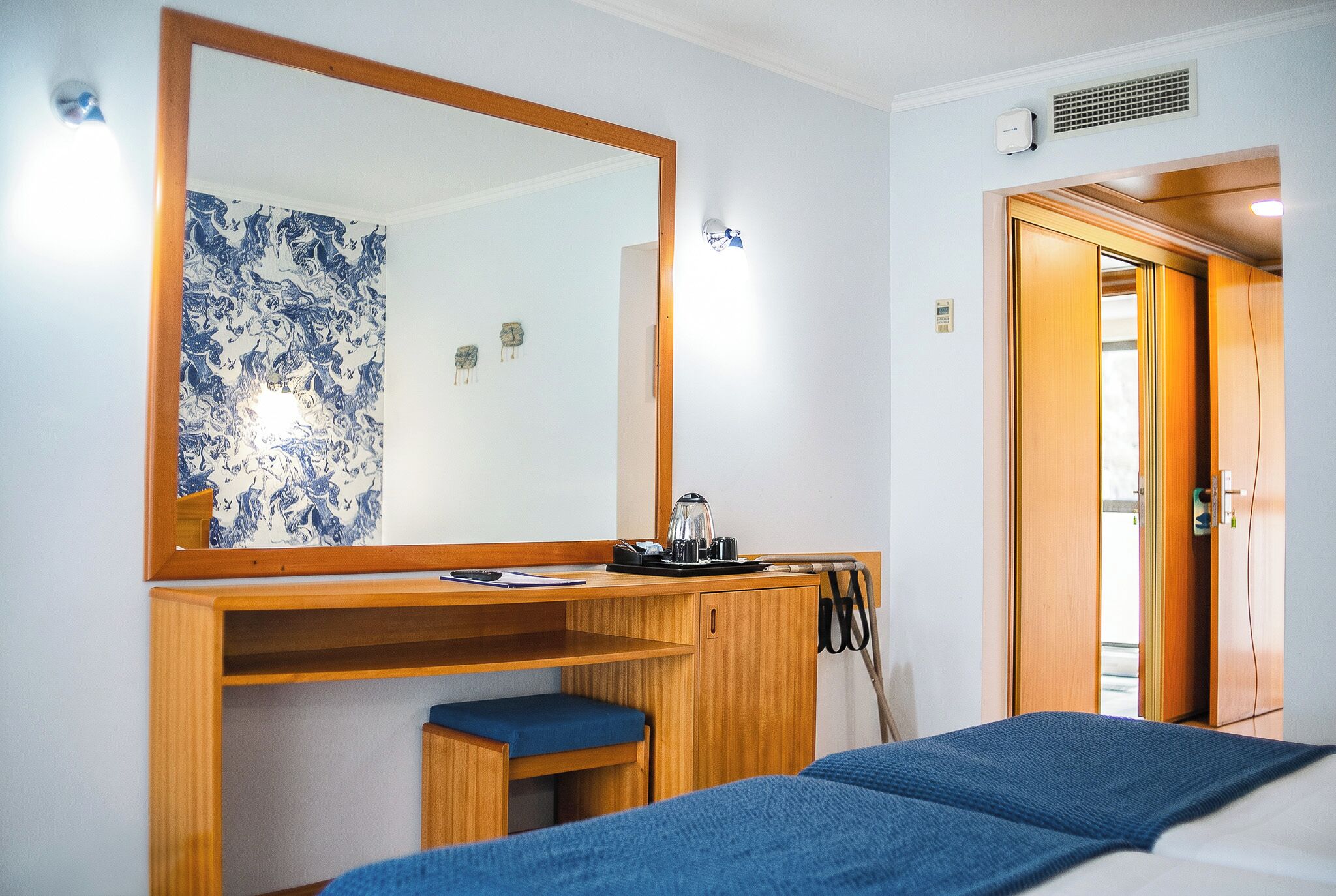 Habitación de hotel con un escritorio de madera, un espejo grande, una cama con manta azul y una puerta que da a otra habitación.