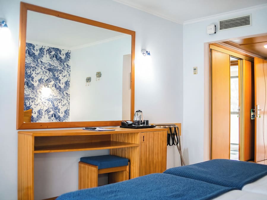 Quarto de hotel com escrivaninha de madeira, espelho grande, cama com manta azul e porta para outro cômodo.