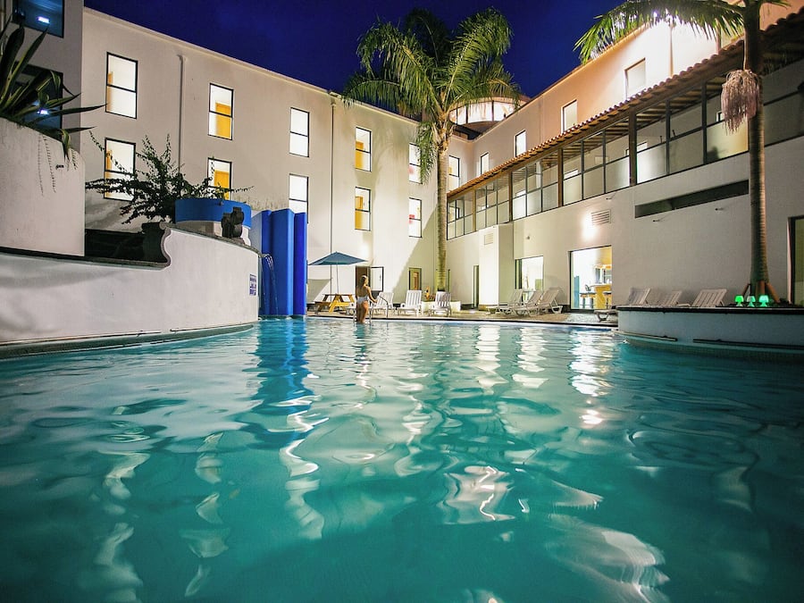 A swimming pool with clear water, surrounded by a building with multiple windows and a palm tree.