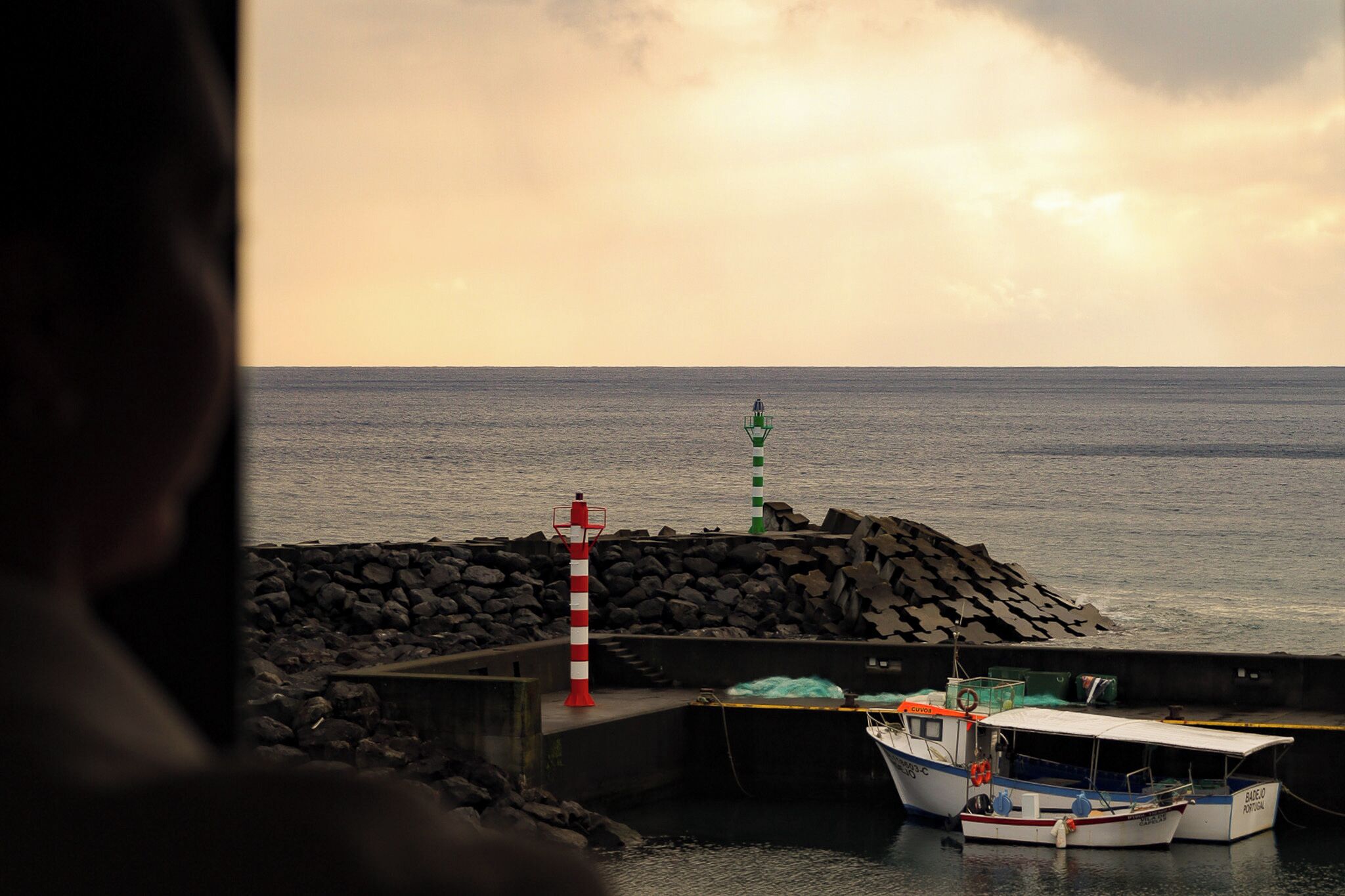 A coastal scene with a lighthouse, a rocky breakwater, and two boats docked at a pier.