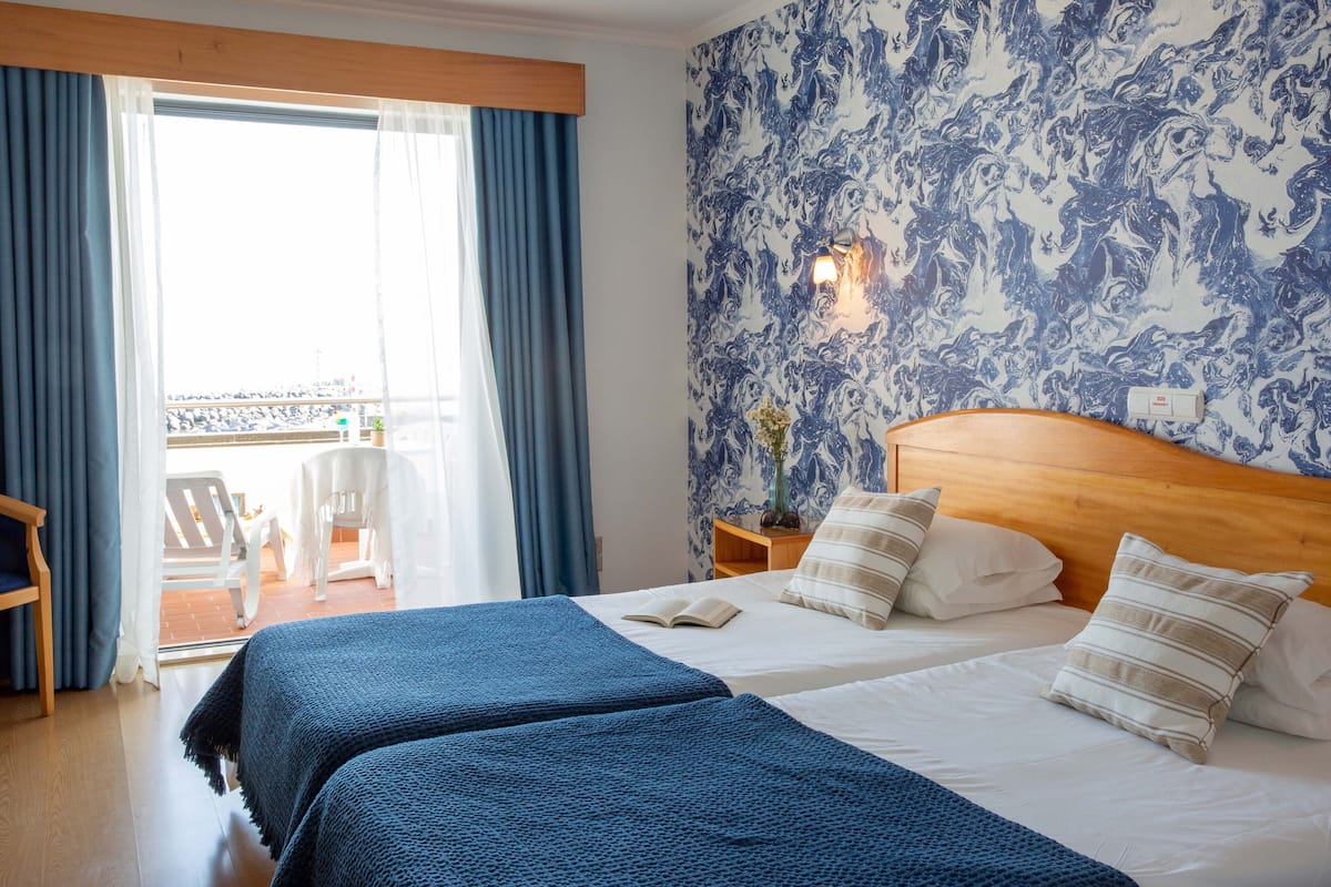 A hotel room with two beds, a wooden headboard, a large window with curtains, and a view of the outside.