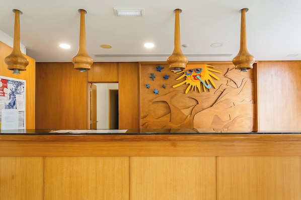 A reception desk with wooden paneling, a wall-mounted sun decoration, and pendant lights. - Hotel do Mar (Povoacao)