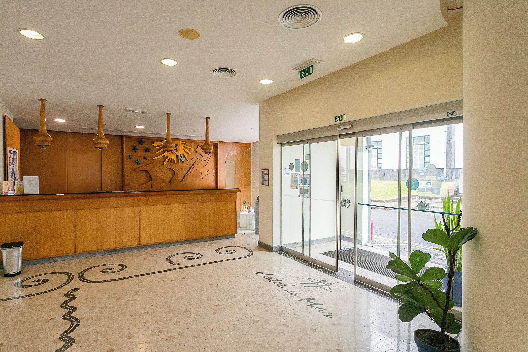 Un área de recepción de hotel con mostrador de madera, puerta de cristal y una planta en maceta.