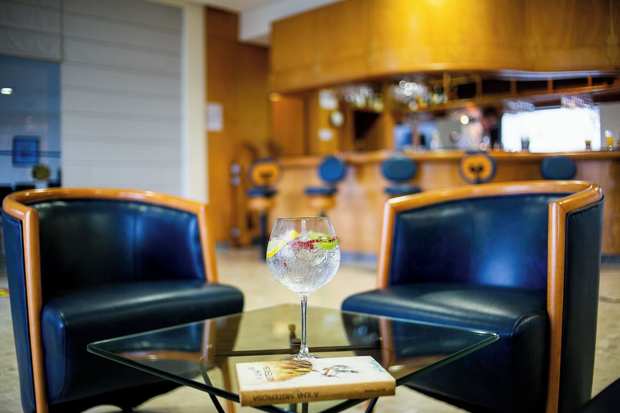 A bar area with two leather chairs, a glass table with a book, and a bar counter with bottles and glasses.