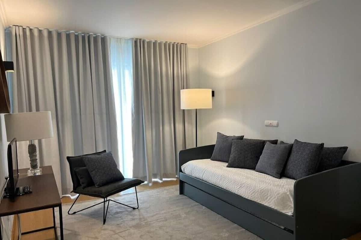 A modern hotel room with a grey sofa, a black chair, a desk with a lamp, and a large window with curtains.
