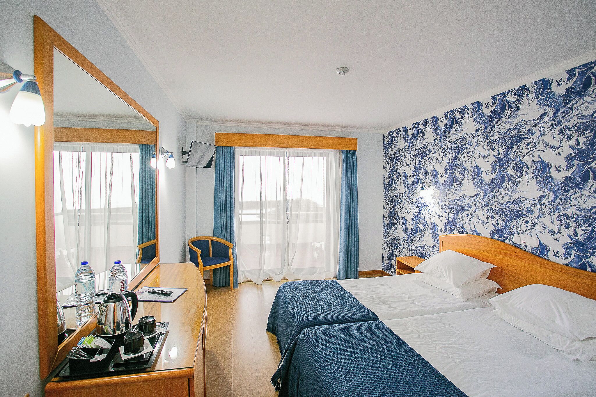 A hotel room with a bed, a desk with a chair, a mirror, and a wall with a blue pattern.