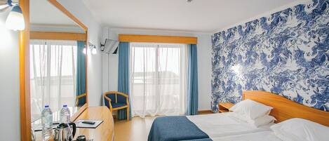 A hotel room with a bed, a desk with a chair, a mirror, and a wall with a blue pattern.
