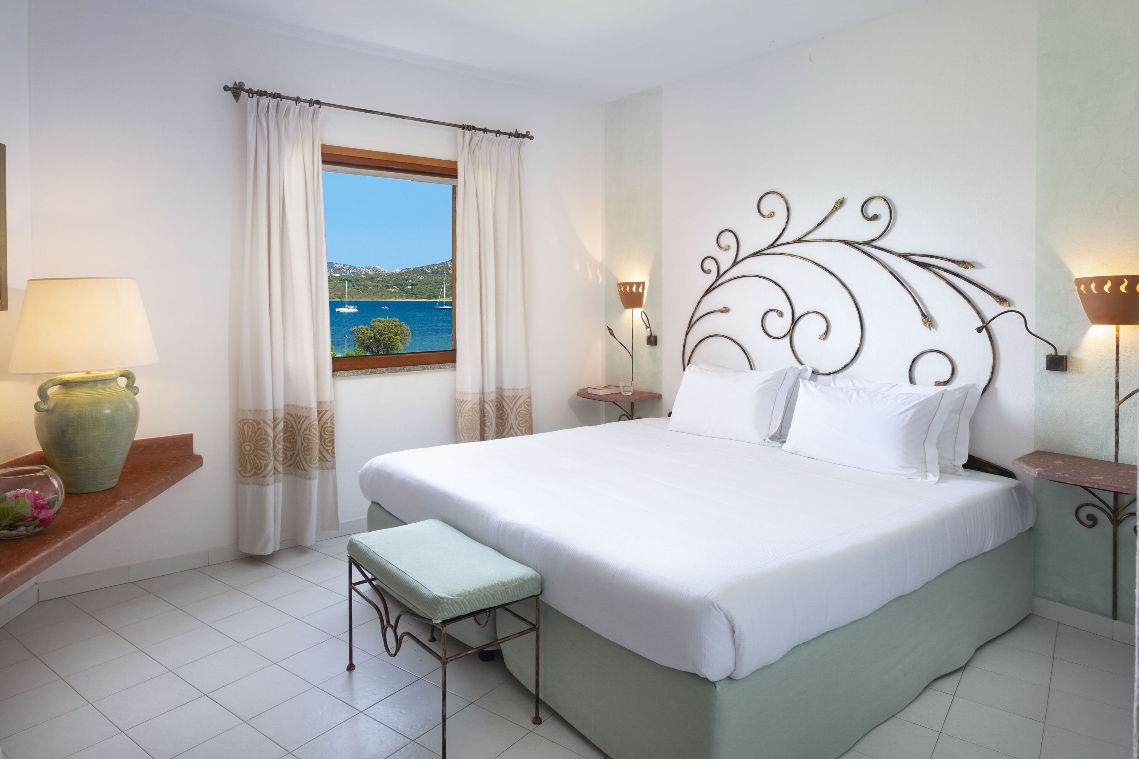 Een slaapkamer met een groot bed, uitzicht op de zee, een vaas met bloemen en een lamp op een nachtkastje.