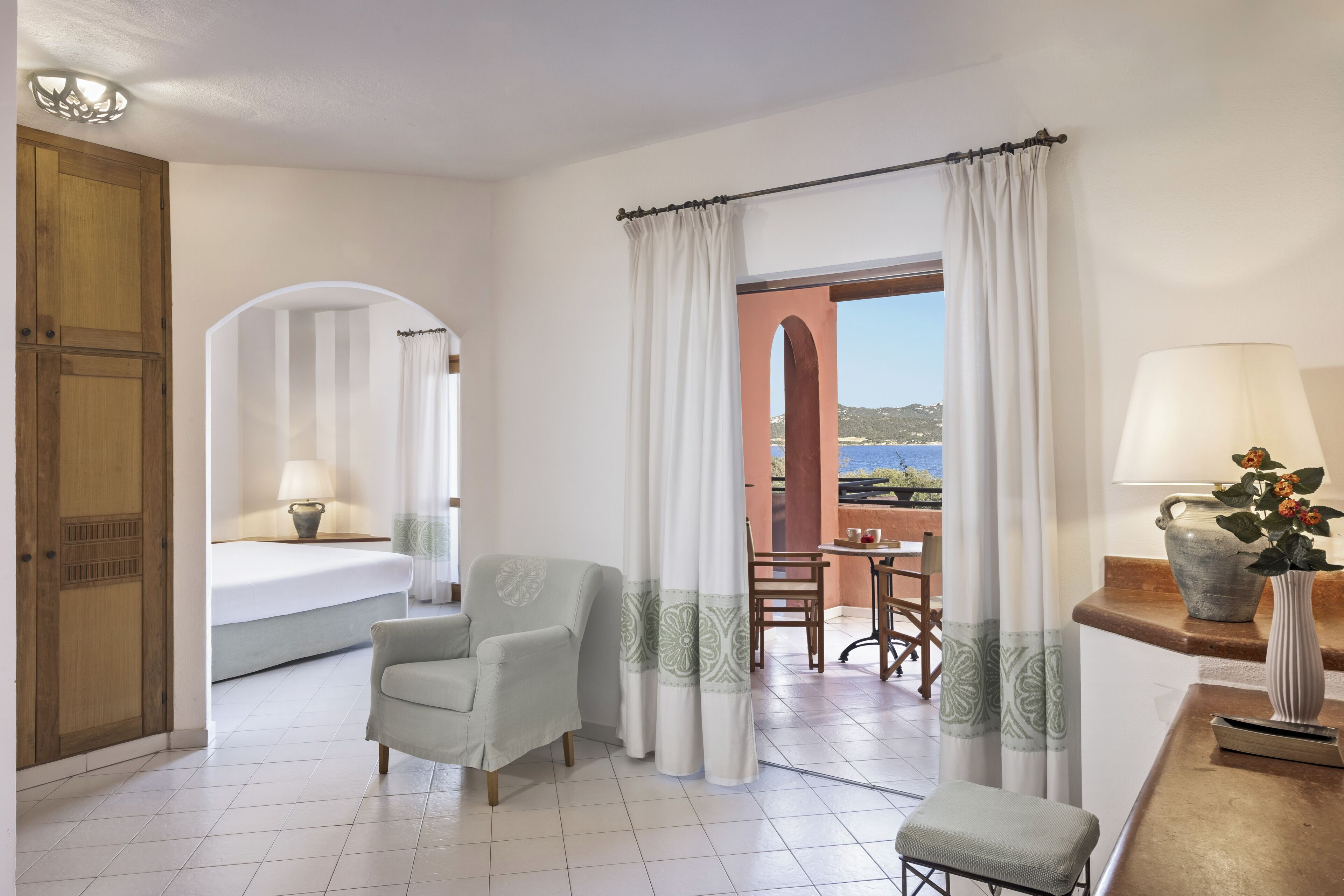Ein Hotelzimmer mit Bett, Stuhl, Tisch, Lampe und Meerblick.
