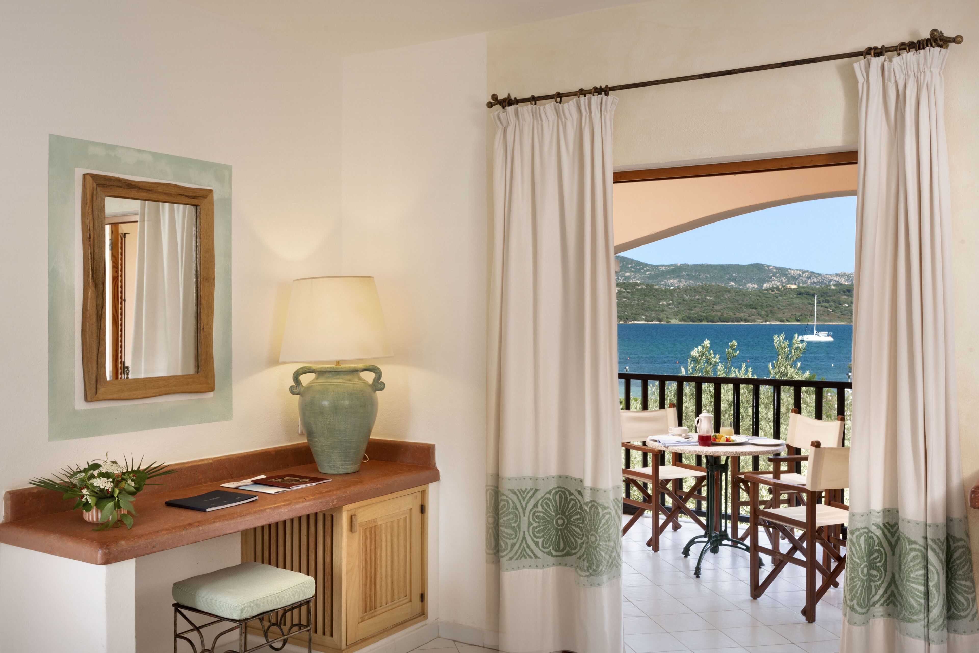A room with a view of the sea, a table with chairs, a lamp, a mirror, and a cabinet.