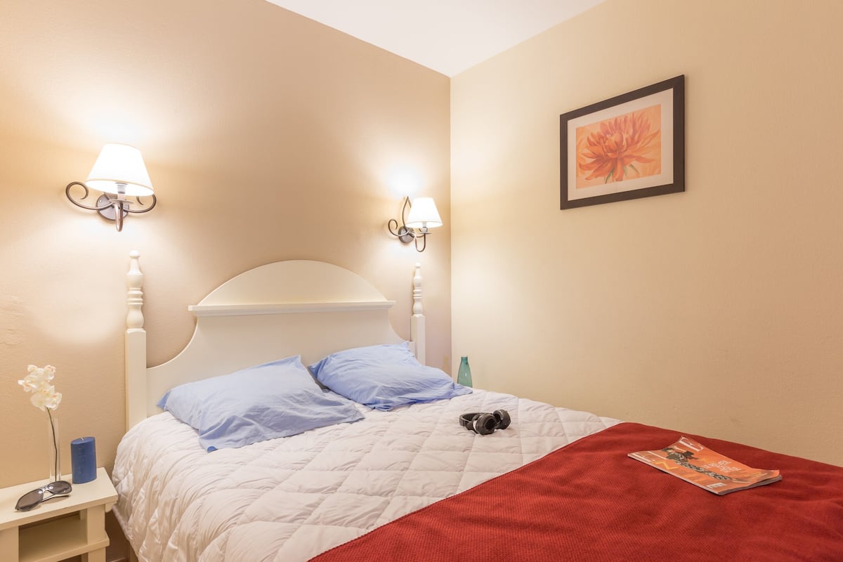Een net opgemaakt bed met een wit hoofdeinde, blauwe kussens en een rode deken. Aan beide zijden van het bed staan wandlampen. Daarboven hangt een fotokader met een bloem.
