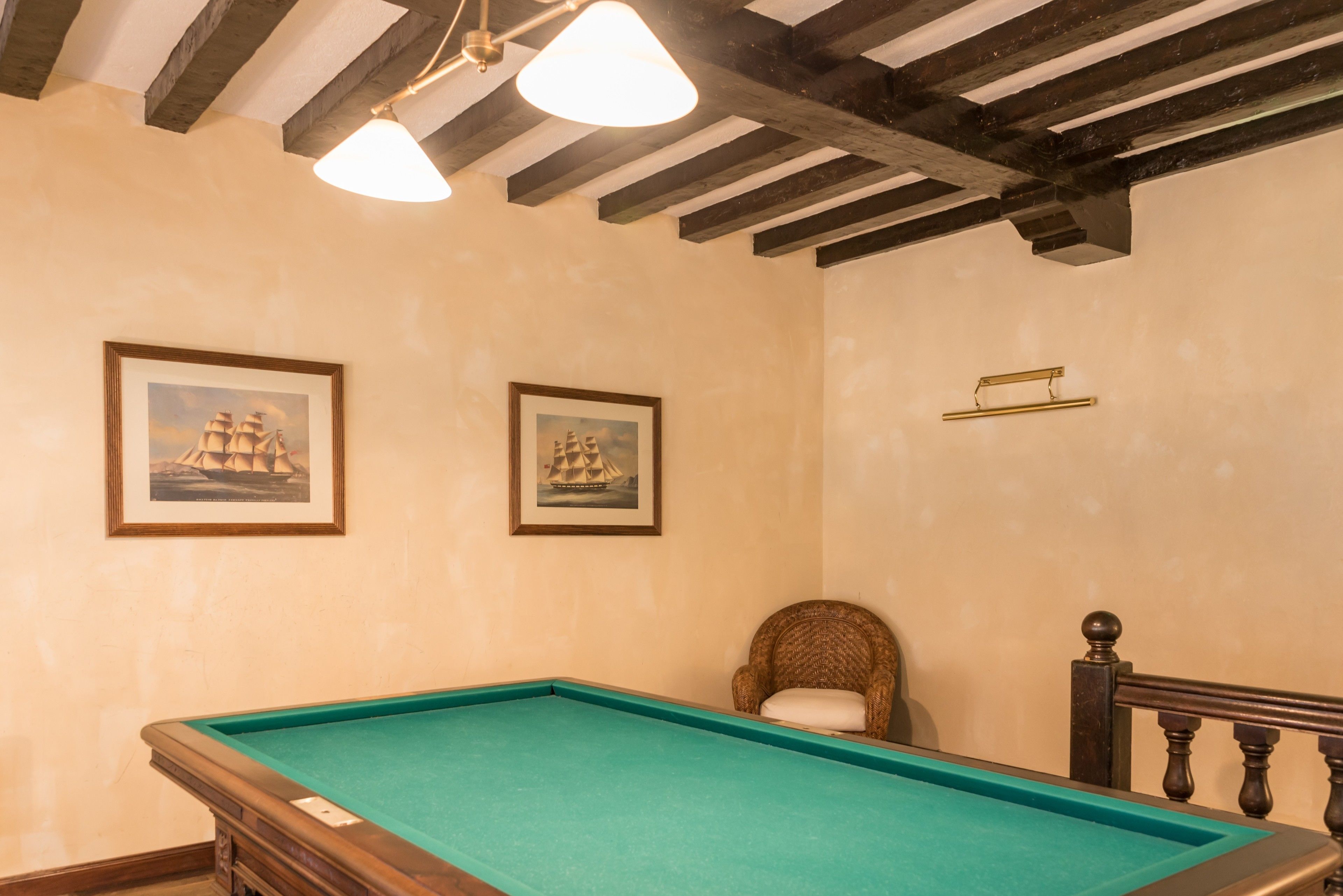Una mesa de billar en una habitación con vigas de madera en el techo, dos cuadros en la pared y una silla de mimbre.