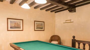 Une table de billard dans une pièce dont le plafond est orné de poutres en bois, avec deux tableaux encadrés au mur et un fauteuil en osier.