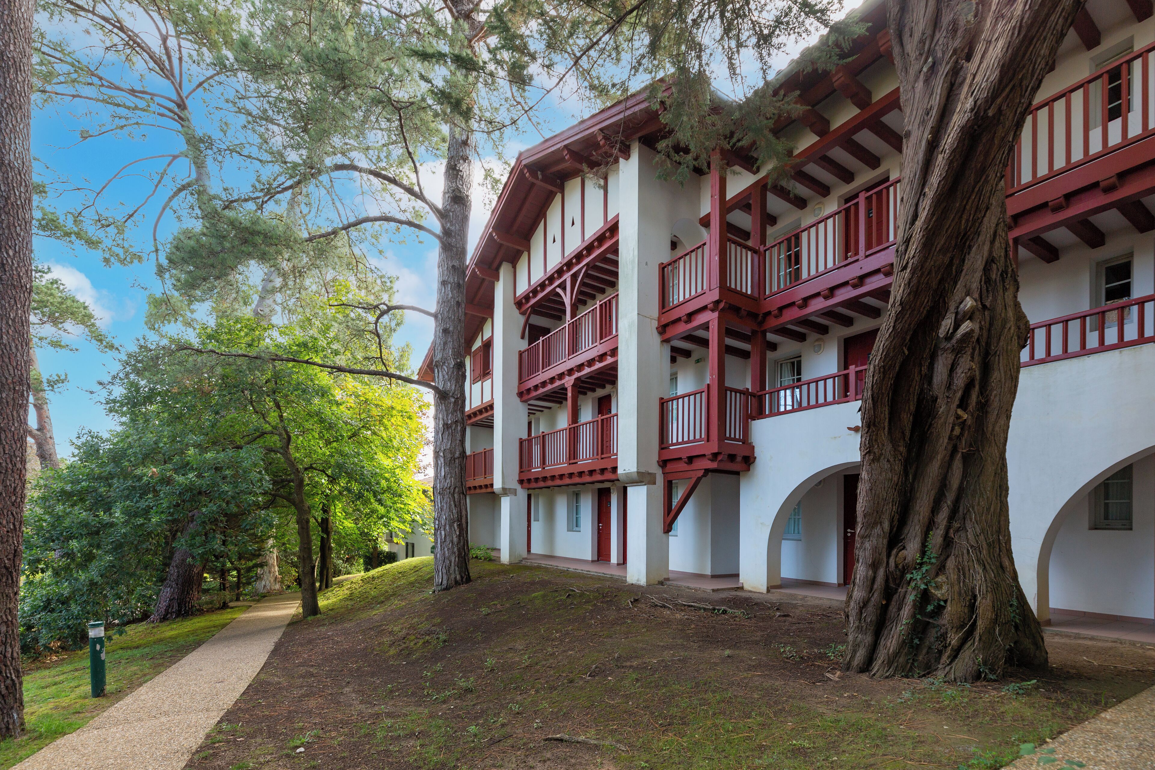 Un edificio con fachada blanca y balcones rojos, rodeado de árboles y un sendero.