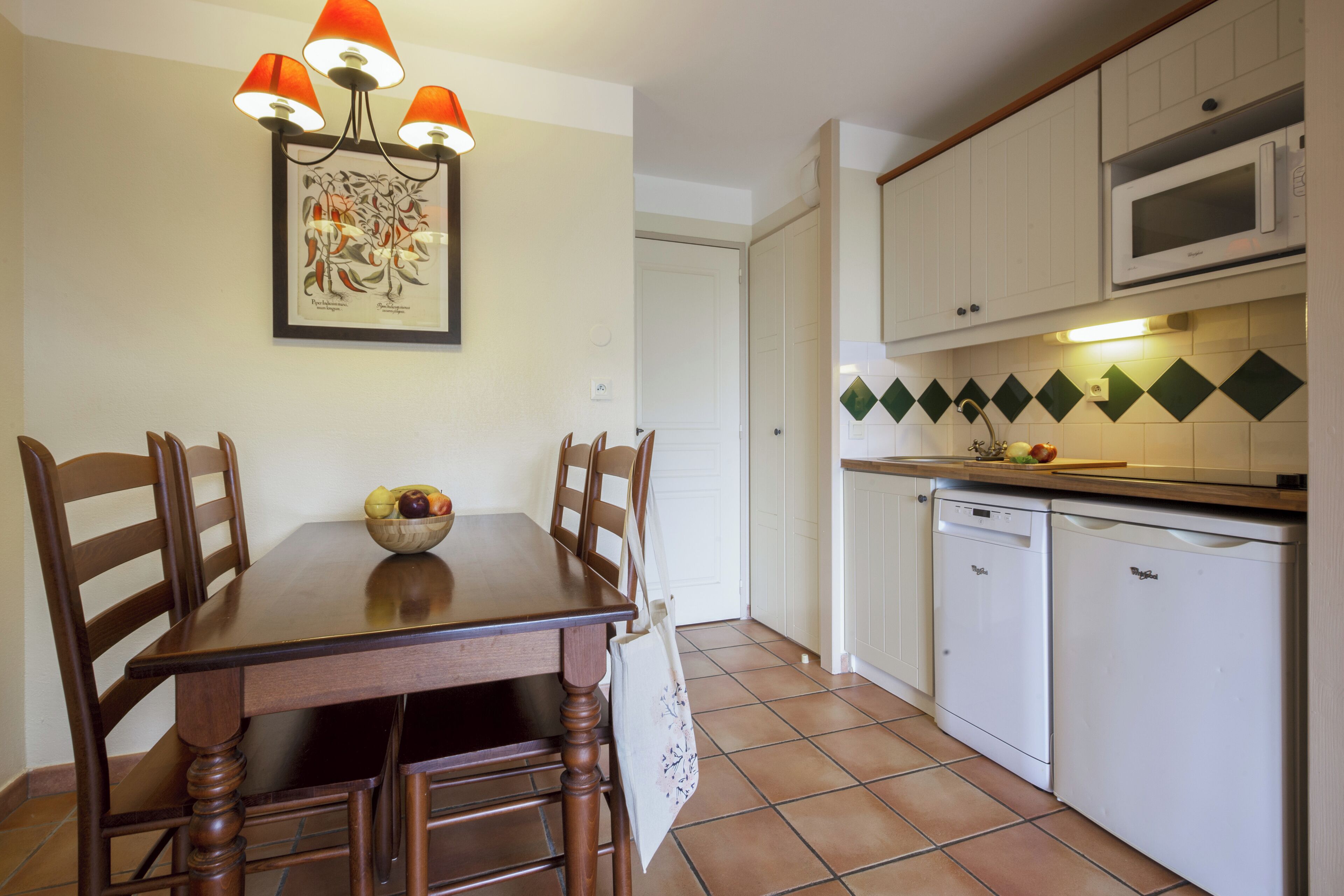 Ein Essbereich mit einem Holztisch, Stühlen und einer Schüssel mit Obst. An der Wand hängt ein Gemälde und es gibt eine Küchenzeile mit weißen Schränken und einer Mikrowelle.