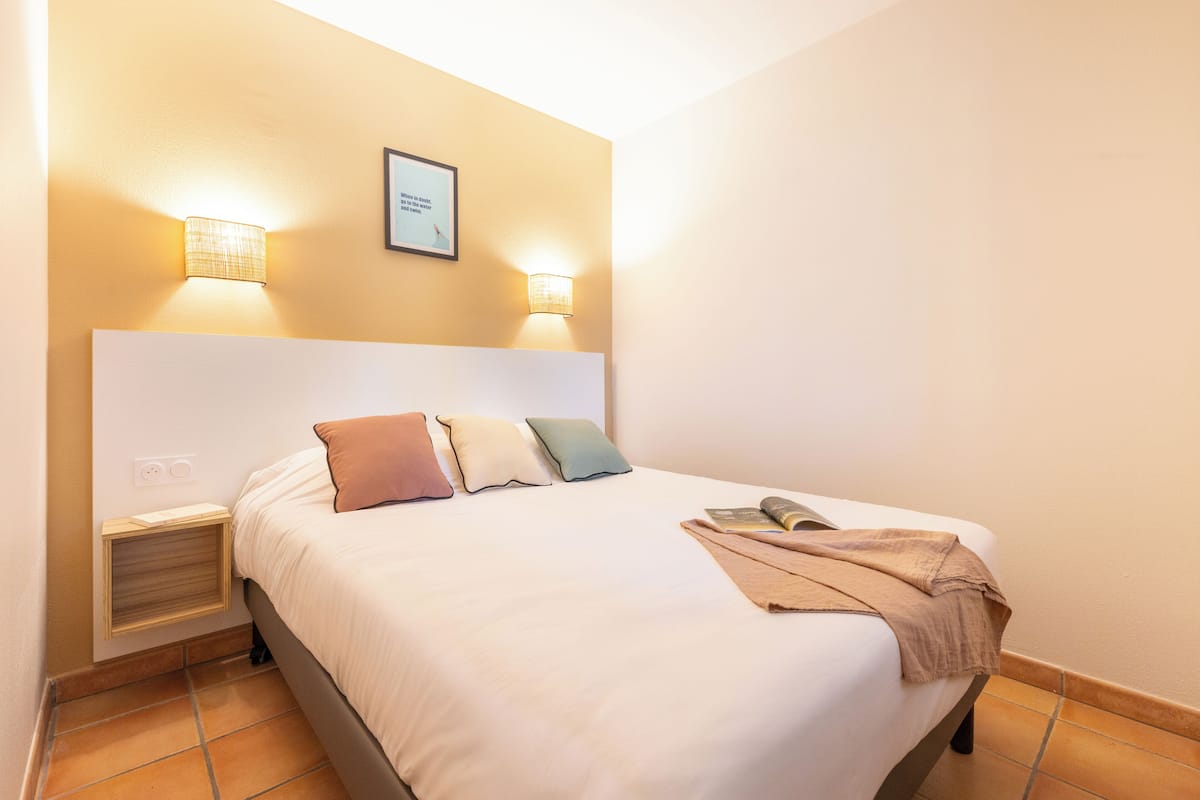 Een opgemaakt bed met kussens en dekbed, een nachtkastje met lamp, en een lijst met een citaat aan de muur.
