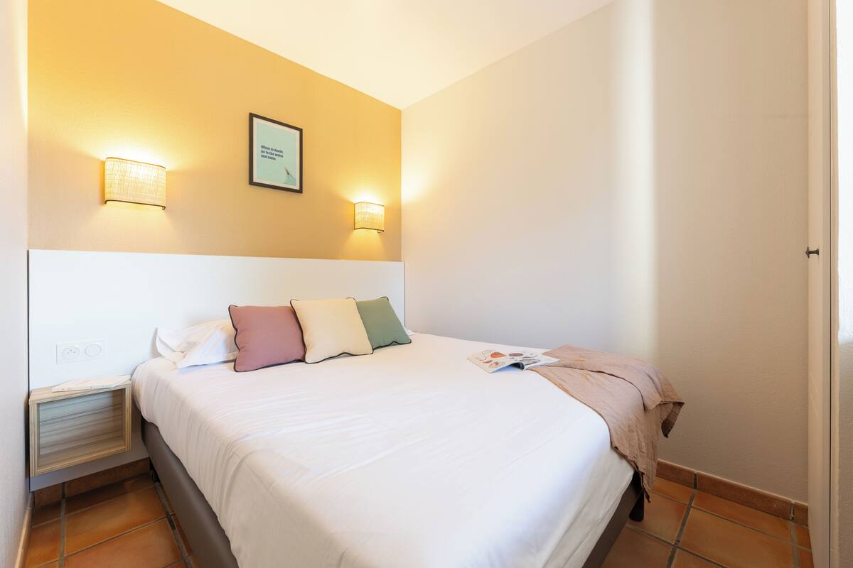 Een net opgemaakt bed met kussens, een nachtkastje met een lamp, en een lijst met een foto aan de muur.