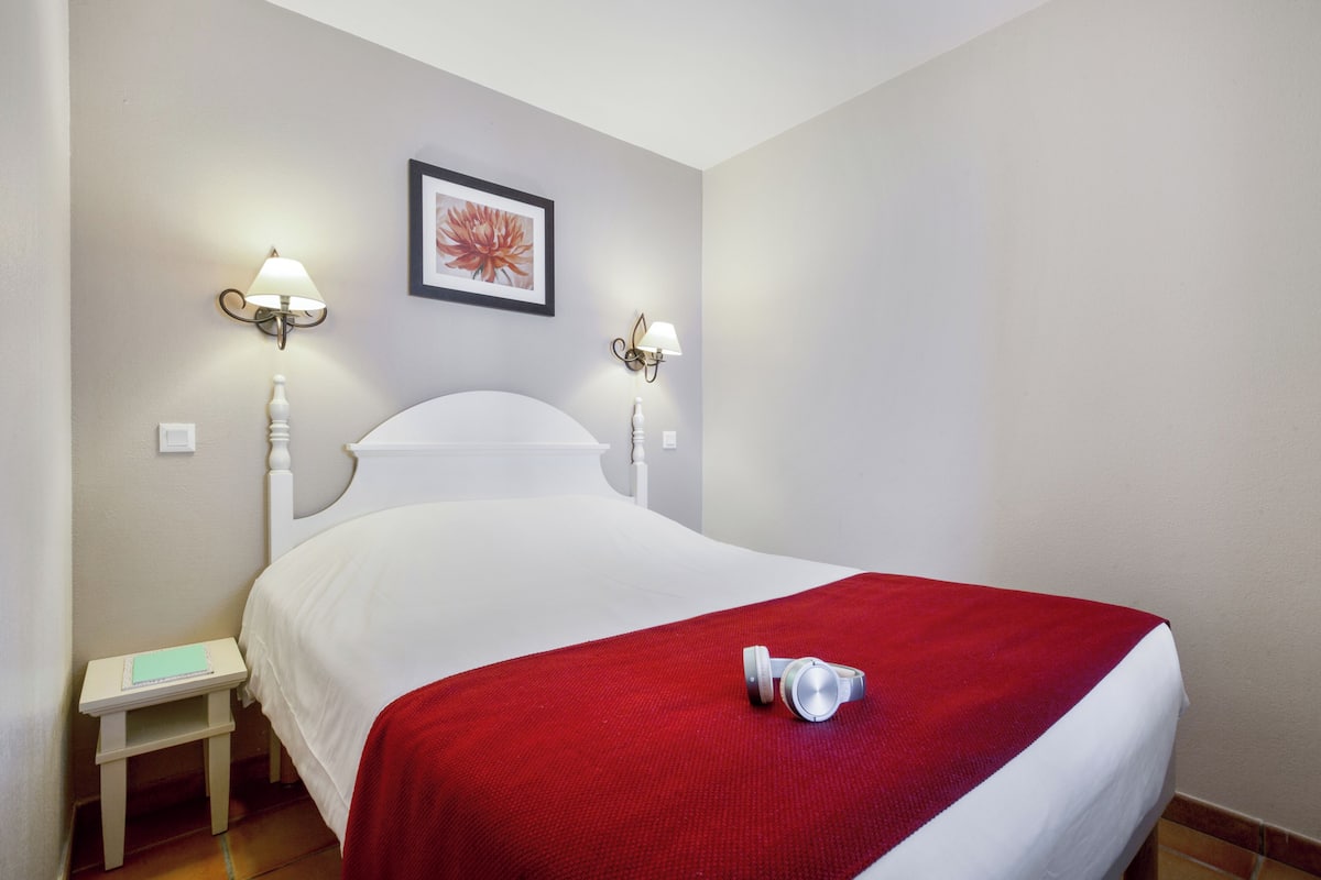 Een net opgemaakt bed met een rode deken en een koptelefoon erop. Aan beide zijden van het bed hangen twee wandlampen.