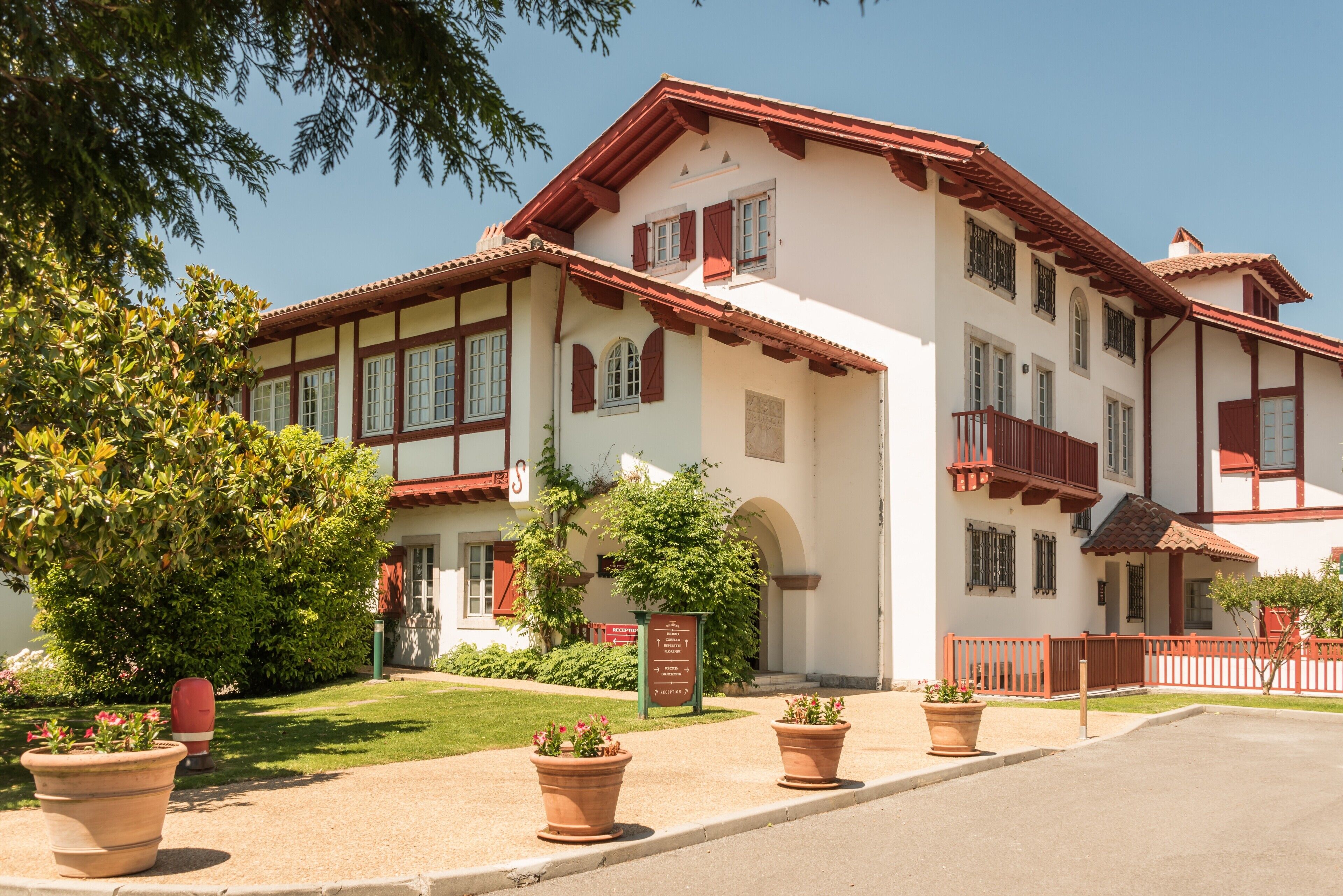 A two-story building with a traditional architectural style, featuring a red-tiled roof and white walls. There are potted plants in the foreground and a signboard in front of the building.