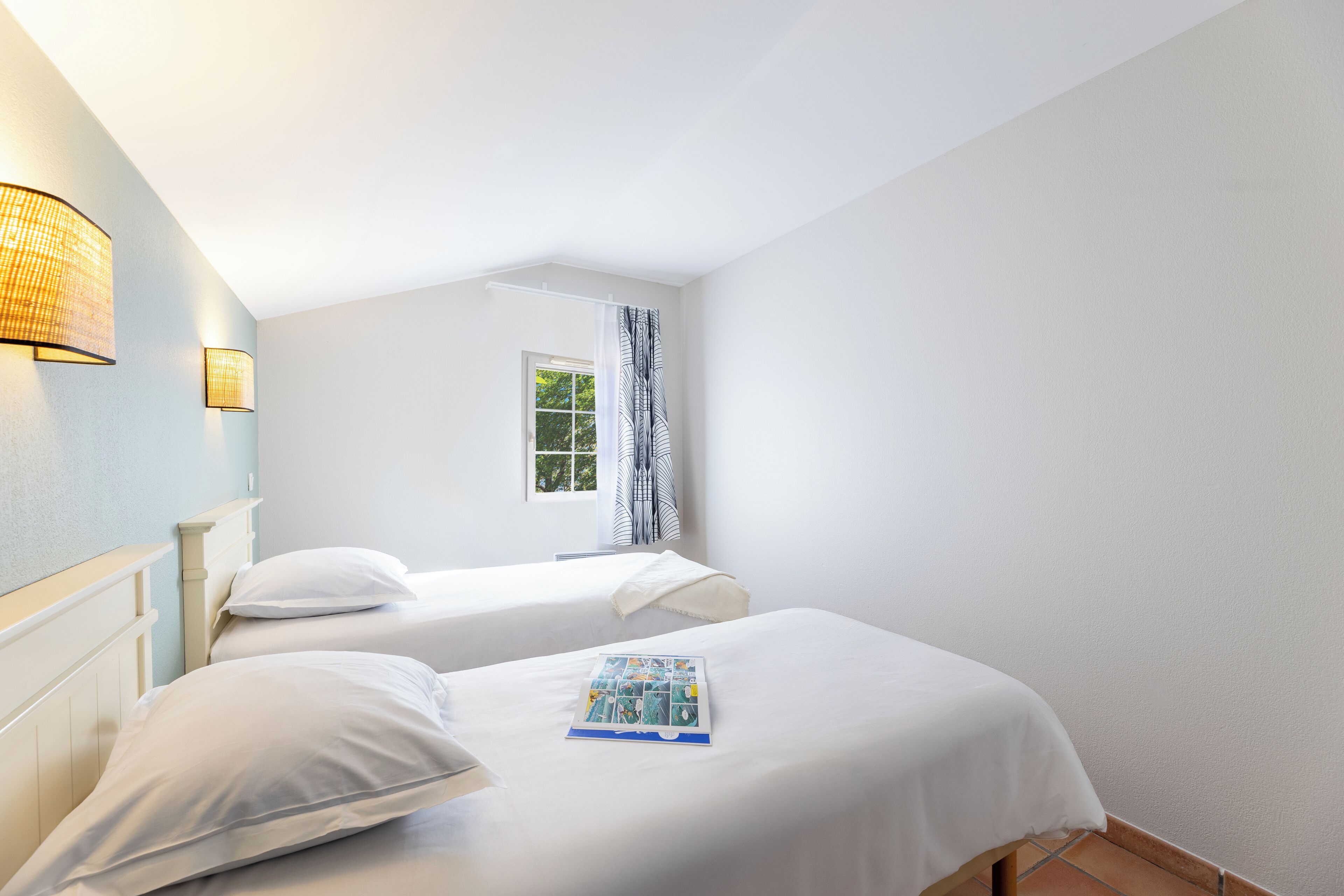 Une chambre d’hôtel avec deux lits simples, une fenêtre donnant sur un paysage agréable et une lampe fixée au mur.
