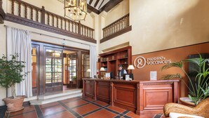 A reception area with a wooden reception desk, a sign for 'Pierre & Vacances', and a check-in counter. There are potted plants and a wicker sofa in the space.