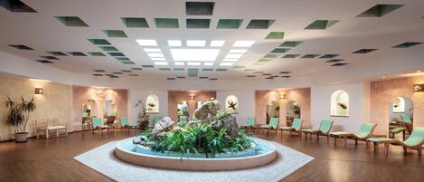 A spacious lobby with a central water feature, wooden flooring, and green chairs arranged around it.