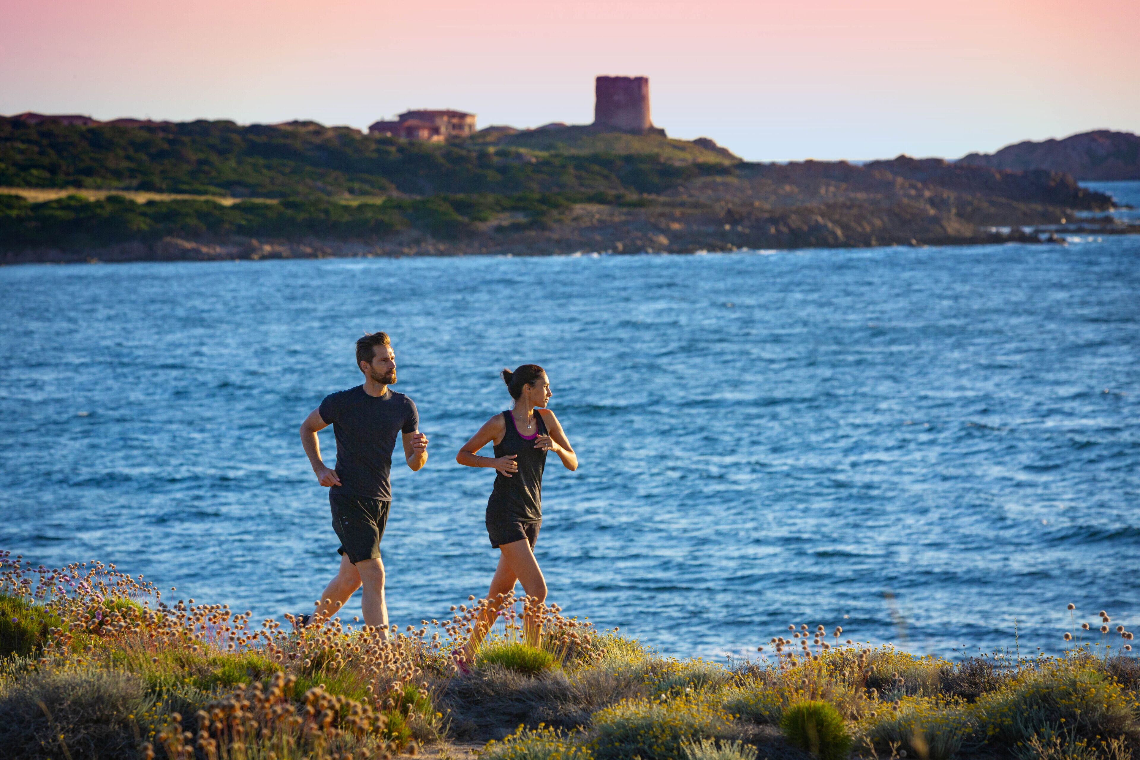 Two people running along a coastal path with a body of water and a distant castle-like structure.
