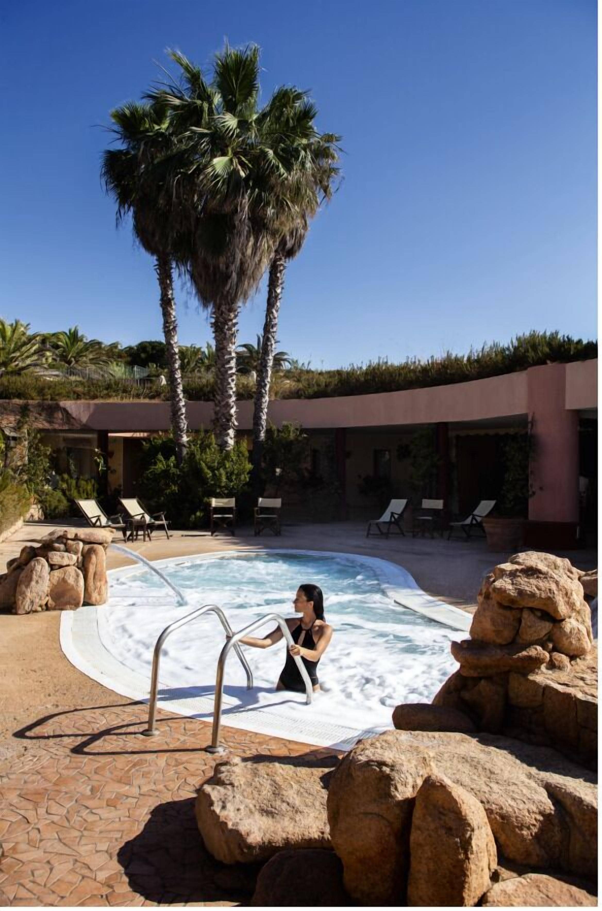 A pool with a hot tub, palm trees, lounge chairs, and a rocky landscape.
