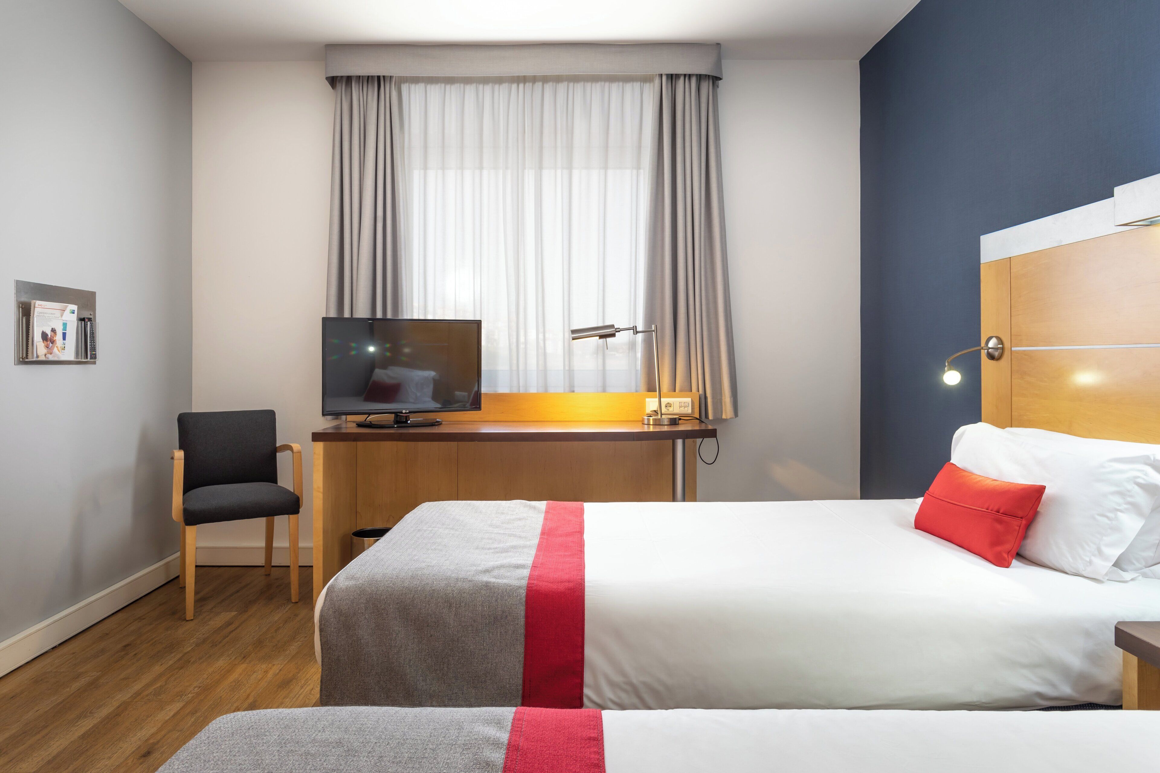 Una habitación de hotel con una cama, un escritorio con televisor, una silla y un ventanal con cortinas.