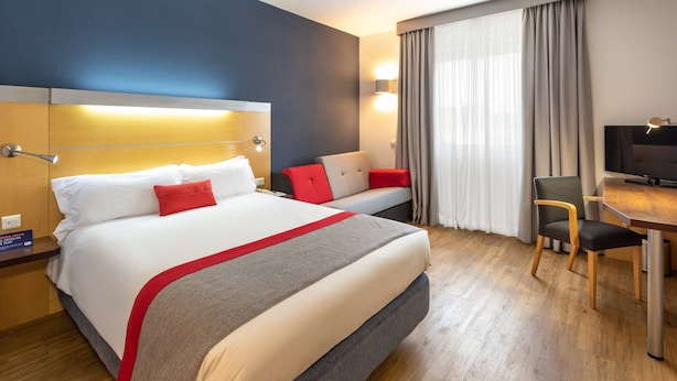 Habitación de hotel con una cama grande, un escritorio, una silla, un televisor y una ventana con cortinas.
