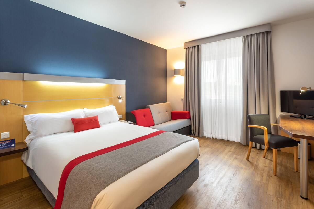 Habitación de hotel con una cama grande, un escritorio, una silla, un televisor y una ventana con cortinas.