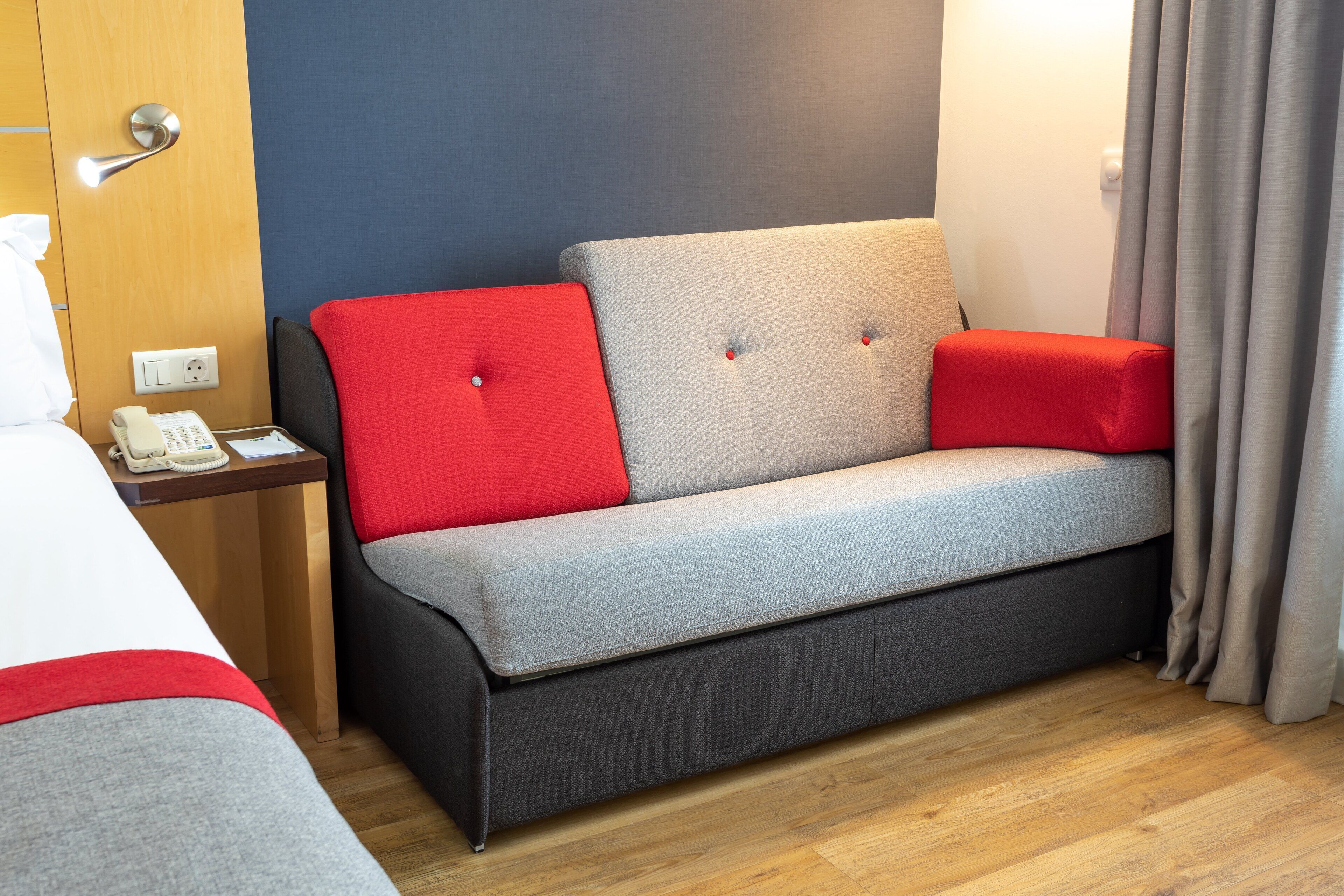 A hotel room with a grey sofa, red cushions, a wooden nightstand with a telephone, and a wooden floor.