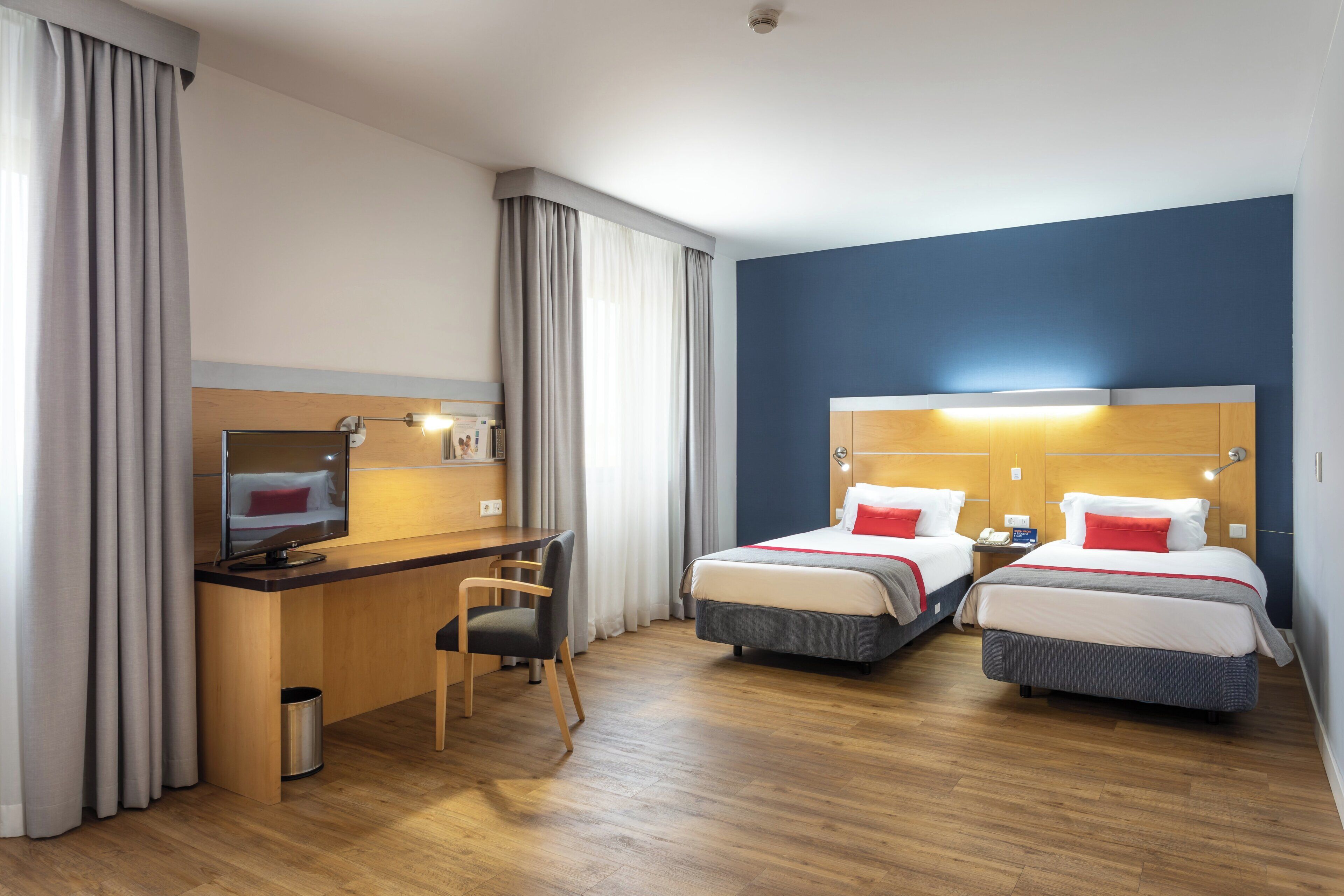 A hotel room with two beds, a desk, a chair, a television, and a trash can.