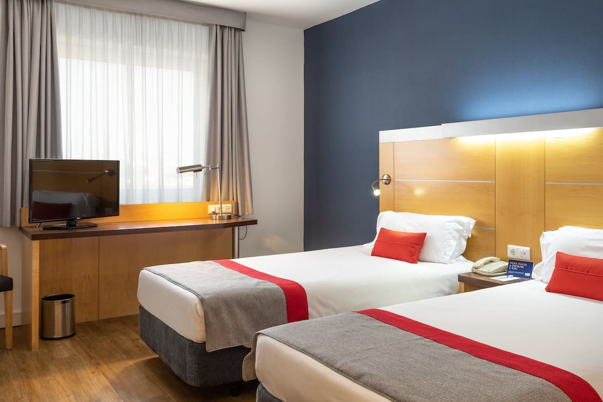 Habitación de hotel con dos camas, un escritorio con computadora, teléfono y ventana con cortinas.