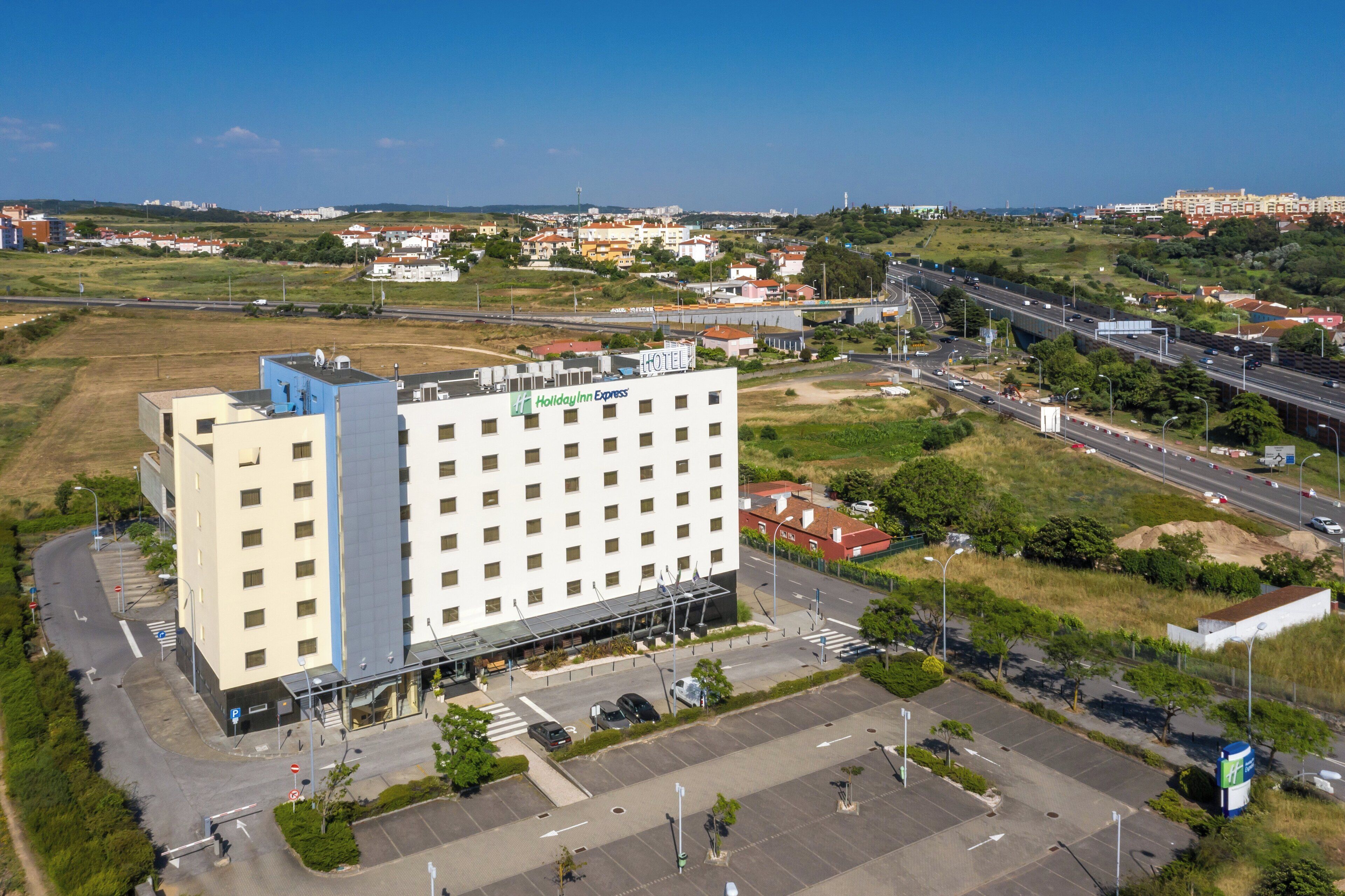 Un hotel de varios pisos con fachada plana, estacionamiento y una autopista al fondo.